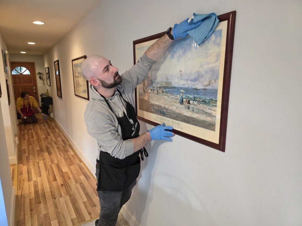 A Rain City Maids cleaner dusting a framed picture in a hallway
