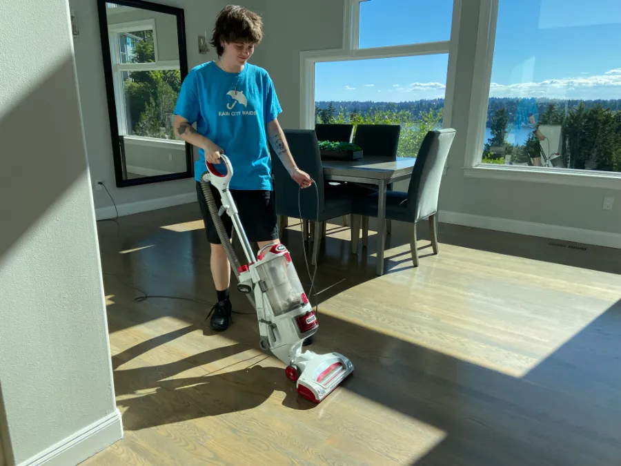 A Rain City Maids cleaner vacuuming beside large lake-view windows